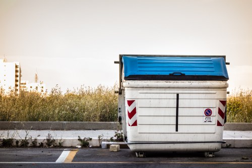 A large, white industrial-sized waste container with a blue lid situated on a paved surface, positioned towards the right side of the image. The container features red and white chevron reflective markings along its sides and a small, circular no-parking sign affixed to its front. The surrounding environment includes a concrete curb in the foreground, with patches of grass and weeds growing alongside it. In the background, there is an open grassy area with low shrubbery and a distant, slightly blurred view of an urban skyline under a clear sky, suggesting a location near Wimbledon or a similar area. The lighting appears natural, with soft, diffused sunlight indicating early morning or late afternoon light, and the overall scene conveys a typical urban or suburban rubbish disposal setting relevant to commercial waste management.
