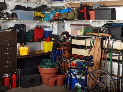 A cluttered storage area or garage containing a mix of household and gardening items, including various plastic storage bins in transparent, black, and grey colors, some filled with objects, placed on wire shelving units. In the foreground, there is a yellow plastic container, a red bag, and a blue box, along with a coiled garden hose in a large terracotta pot and smaller empty pots nearby. To the right, a blue bicycle leans against a wooden shelving unit, which holds a small electronic device with a black cord, a wooden chair, and other miscellaneous items. The background features more shelves stocked with tools, boxes, and outdoor equipment, with some bundles of garden tools hanging or stacked. The environment appears to be a typical domestic garage or storage space, with neatly arranged objects and a dense collection of household, garden, and maintenance supplies, consistent with an area where rubbish might accumulate before removal by a professional waste removal service in Wimbledon.