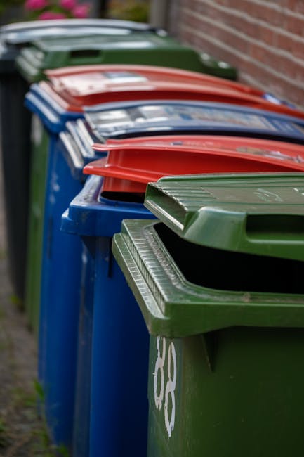 A collection of overflowing rubbish bins and scattered waste on a paved urban sidewalk near a parking area. The waste includes black, red, and grey bins, with black and grey ones appearing to be full,